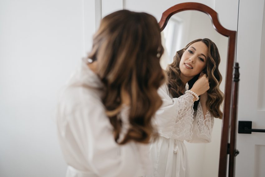 Bride admiring her lace wedding gown hanging by a bright window, capturing a serene and intimate bridal preparation moment.